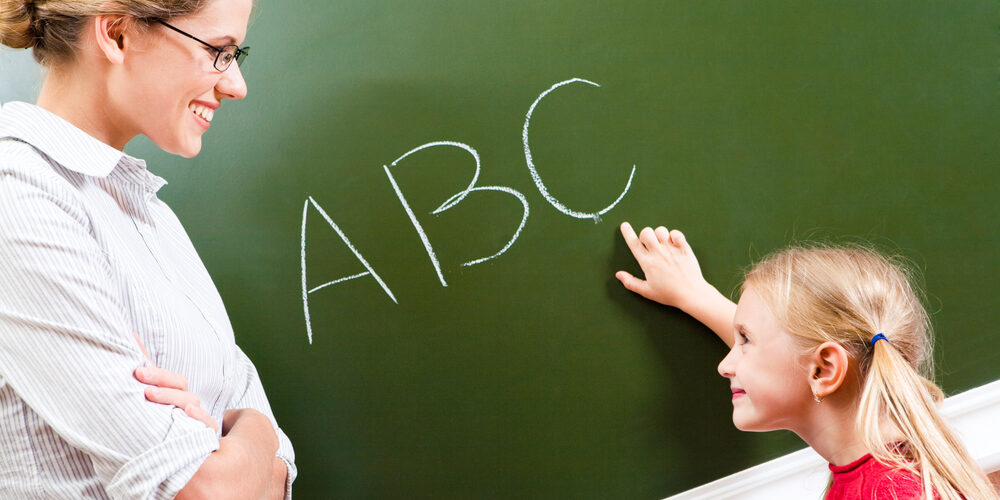Image of smart girl pointing at letter on blackboard and looking at her teacher with smile